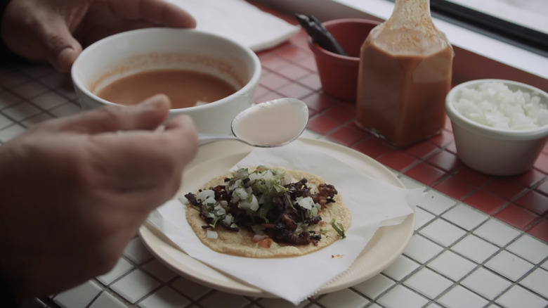 A hand constructing a birria taco with consomme and ingredients in the back.