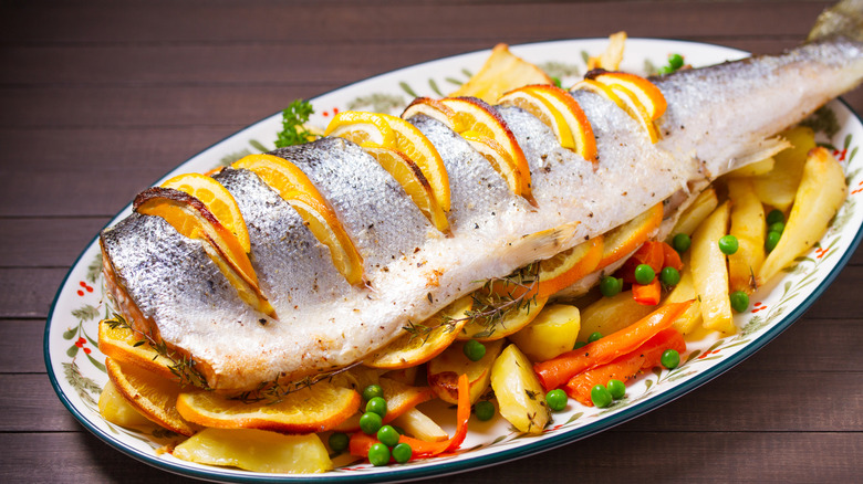 Whole roasted salmon skin-on with lemon wedges on a bed of potatoes, peas, and carrots