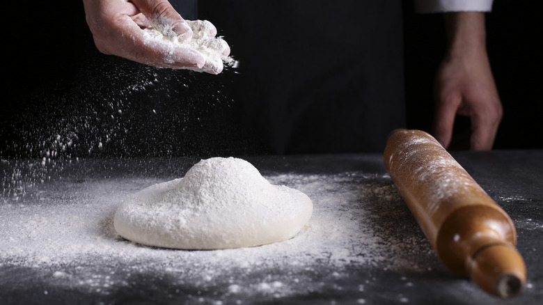 Person's hand sprinkling flour on dough with rolling pin to the right