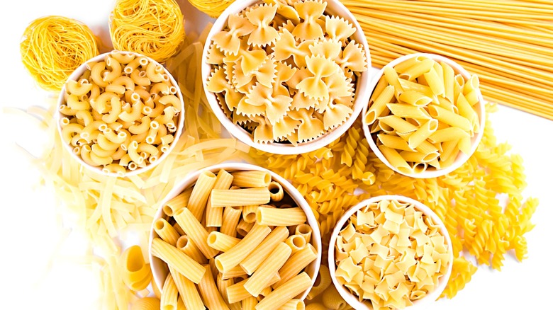 Various dried pasta shapes in white bowls and on white surface