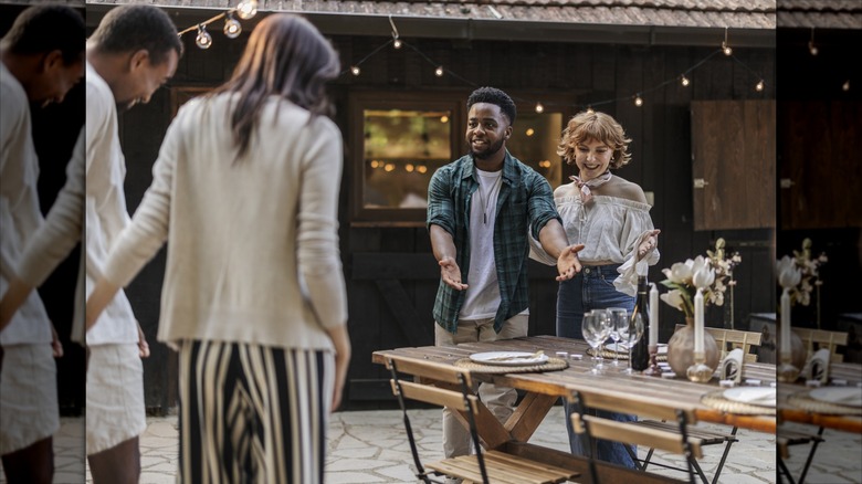 Two friends joyfully greet guests at an outdoor dinner party, pointing to a beautifully set table on a cozy patio