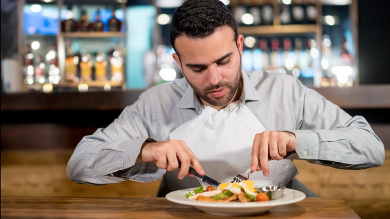 Man cuts chicken salad with fork and knife at restaurant, wearing a napkin in his shirt