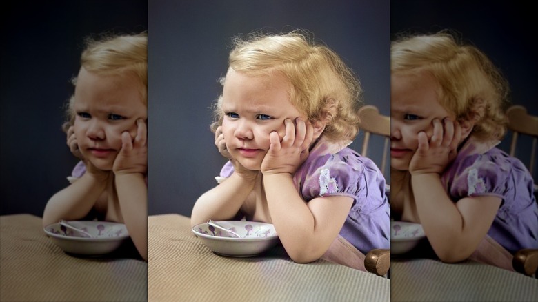 little girl looking defiant, with her elbows on table over small bowl and spoon