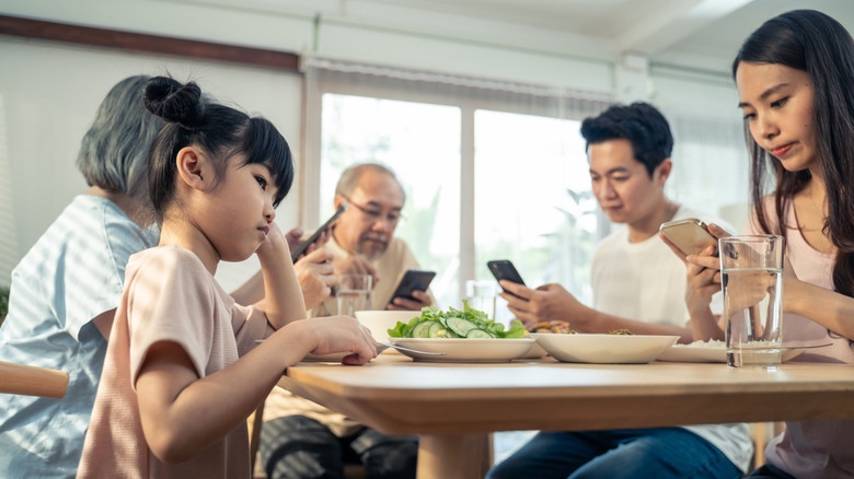 Grandparents and parents sitting at table looking at phones as young daughter ignores plate of salad and looks off bored