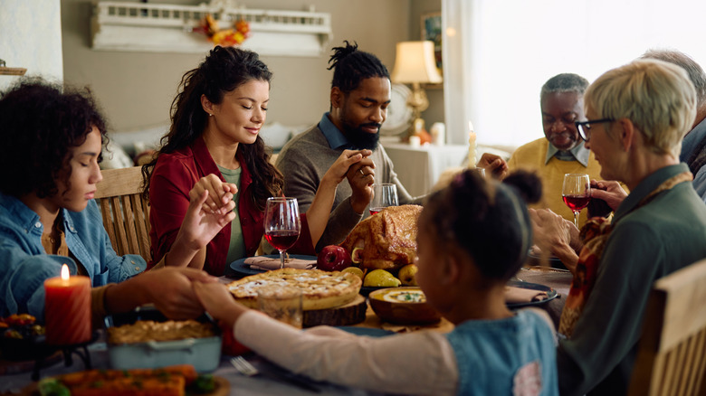 Multi-generational and diverse family hold hands and close eyes to say grace around thanksgiving meal at dining room table