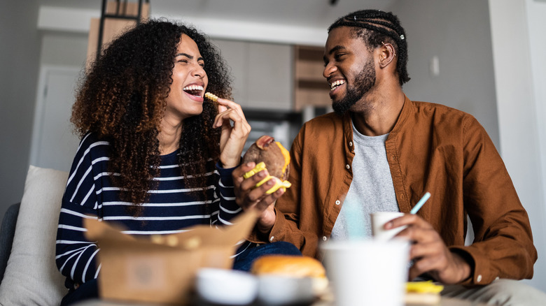 Young couple laugh and share takeout food on sofa in apartment
