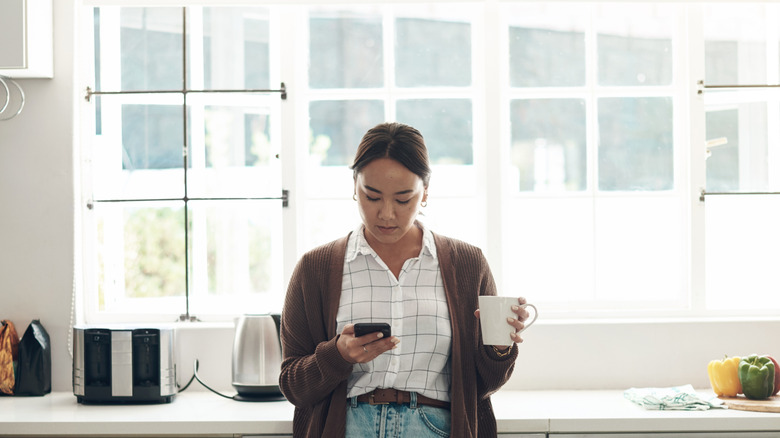 Young woman holding a mug and scrolling on her phone in the kitchen