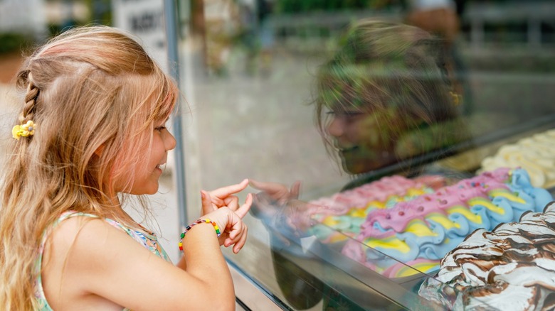 Young girl looking through glass window at a store with different sorts of ice creams and sweets.