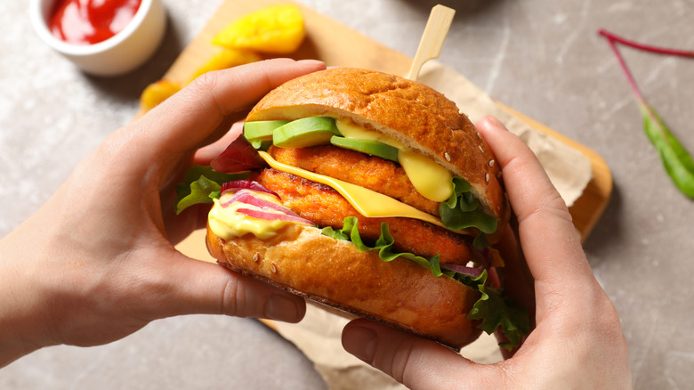 Two hands holding veggie burger with to patties, cheese, avocado, lettuce, and sauce