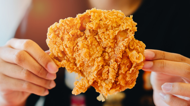 Hands holding a large piece of fried chicken highlighting its crunchy texture.
