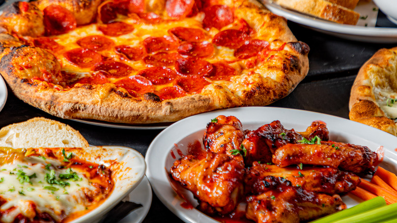Wide shot of pepperoni pizza, chicken wings, and baked pasta.