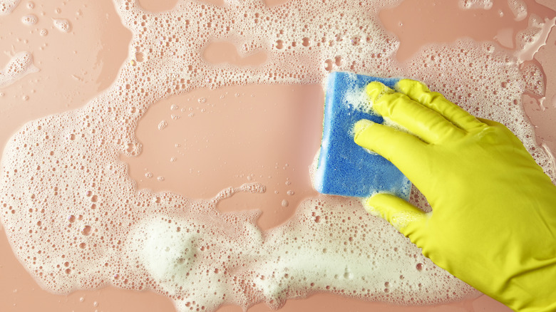 A rubber-gloved hand cleans a pink surface with a sponge and soapy water