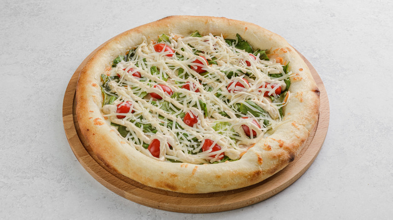 A caesar salad pizza with cherry tomatoes, lettuce, shredded cheese, and dressing on a wood plate