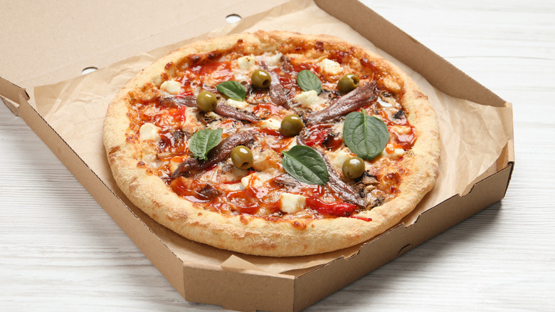 An opened pizza box revealing a pizza topped with green olives, anchovy filets, and fresh spinach leaves