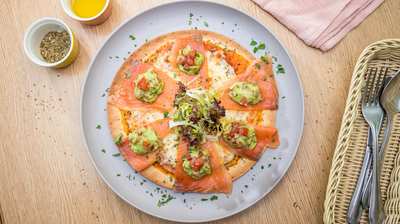 Overhead shot of smoked salmon pizza garnished with salad greens, on a plate surrounded by cutlery and ingredients