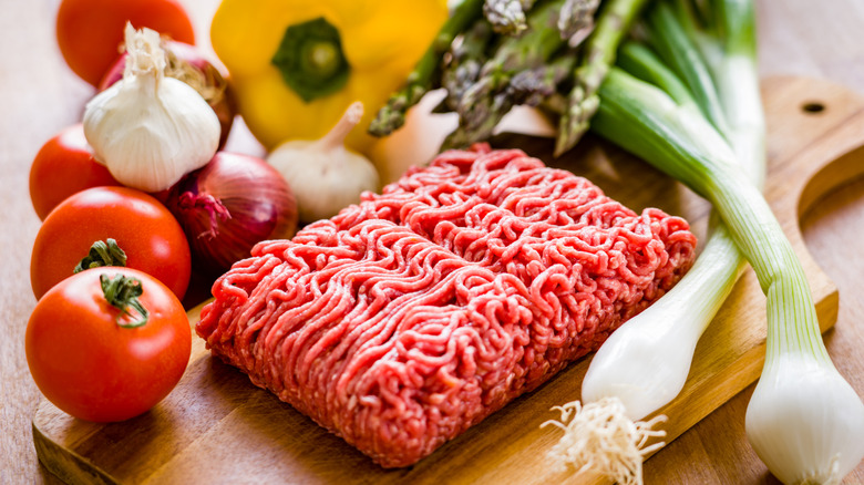 Raw ground beef on wooden cutting board surrounded by vegetables like green onions, tomatoes, and garlic bulbs