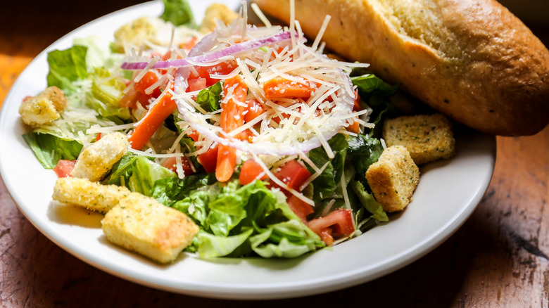 White plate holding salad with croutons, lettuce, tomato, red onion, and cheese with breadstick on the side
