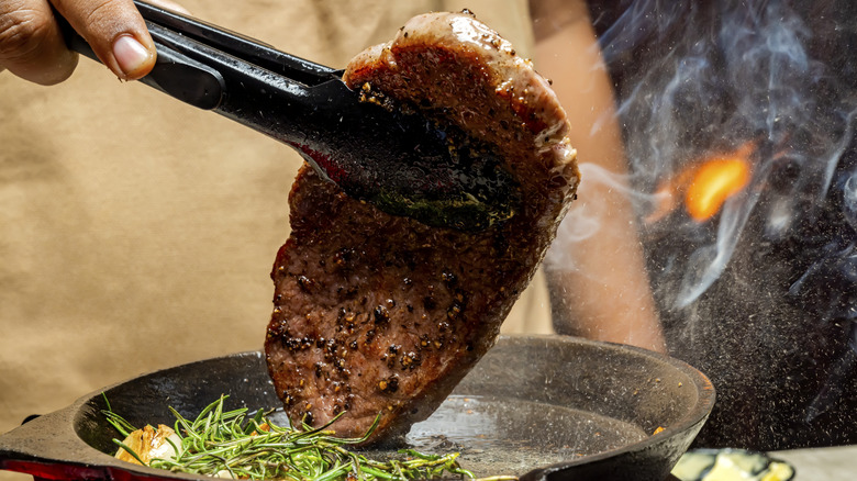 A person holds a steak with tongs above a hot pan with herbs in it.