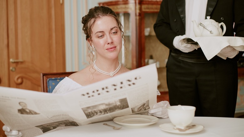 young adult woman reading newspaper at table while  waiter in tuxedo serving tea on tray