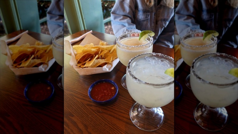 Bowl of tortilla chips and tub of salsa next to two cocktails
