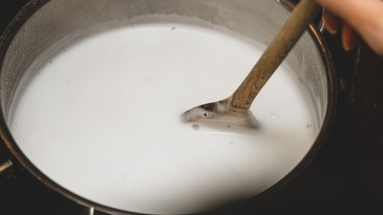 A hand stirrs milk in a pot with a wooden spoon