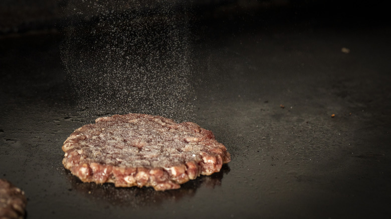 A burger patty being salted as it cooks on a flat griddle
