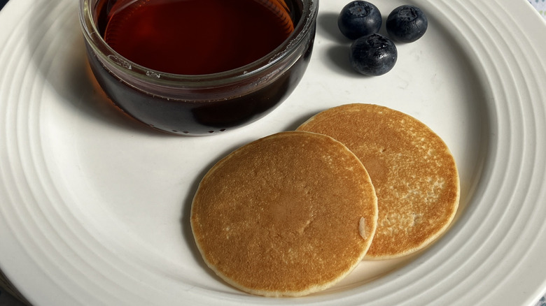 White plate with silver dollar pancakes, glass bowl of syrup, and blueberries