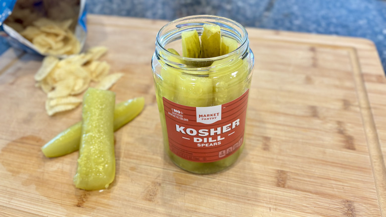 Open glass jar of Market Pantry kosher dill pickle spears and loose spears on a wooden cutting board with potato chips near by