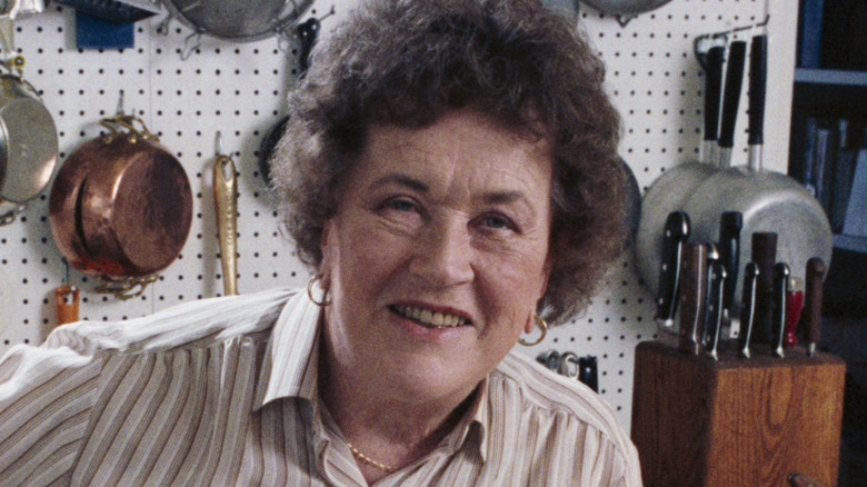 Julia Child poses for the camera in her Santa Barbara kitchen, with several utensils dangling from a pegboard wall behind her.