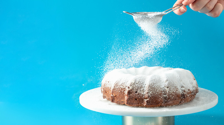 Hand holding small sifter over bundt cake, sifting sugar vigorously.