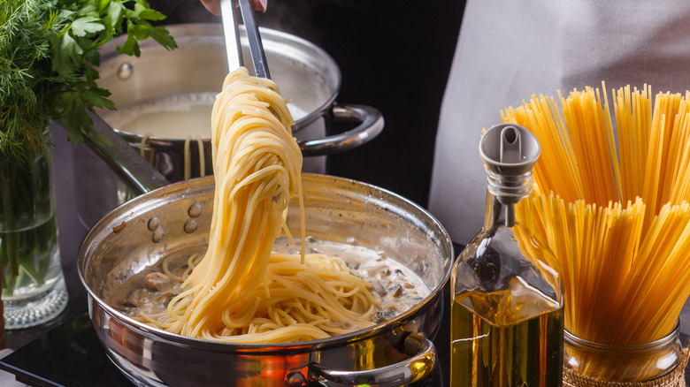 Cooked pasta being removed from a pot with tongs, with oil and dried spaghetti beside it
