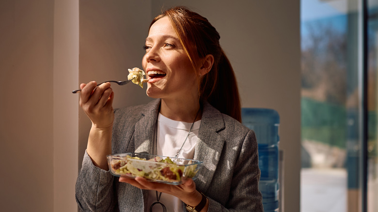 Woman in work attire taking bite of lunch from glass container