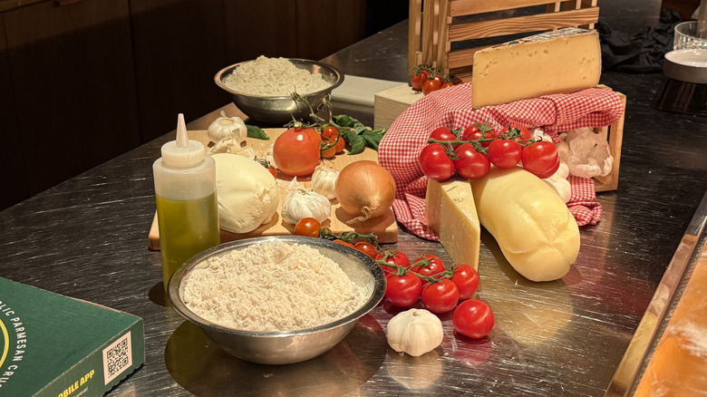 various ingredients that make up Papa Johns' Pan Pizza, including a metal bowl of flour, a plastic bottle of oil, cheeses, oil, garlic, and tomatoes on metal counter