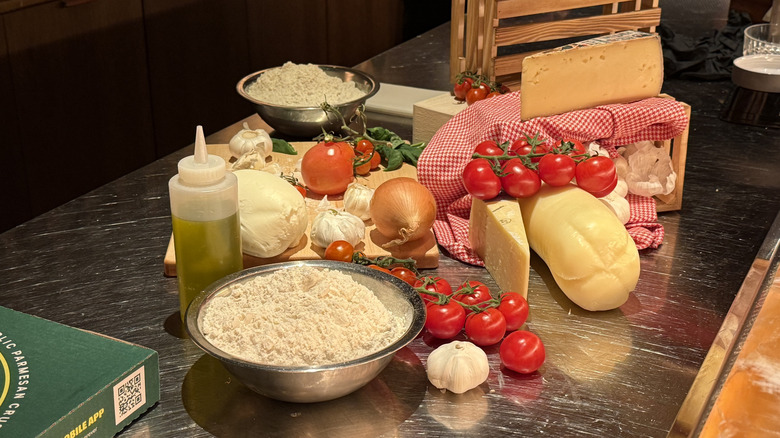 various ingredients that make up Papa Johns' Pan Pizza, including a metal bowl of flour, a plastic bottle of oil, cheeses, oil, garlic, and tomatoes on metal counter