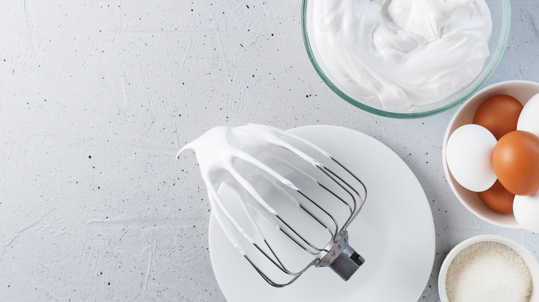 A whisk with and bowl of meringue alongside eggs and sugar