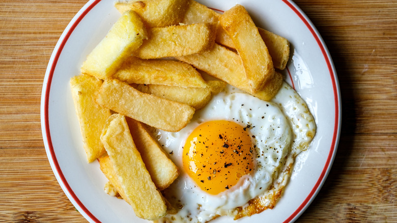 Sunny-side-up egg served with chips on a white plate with red outlines, placed on top of a wooden table.