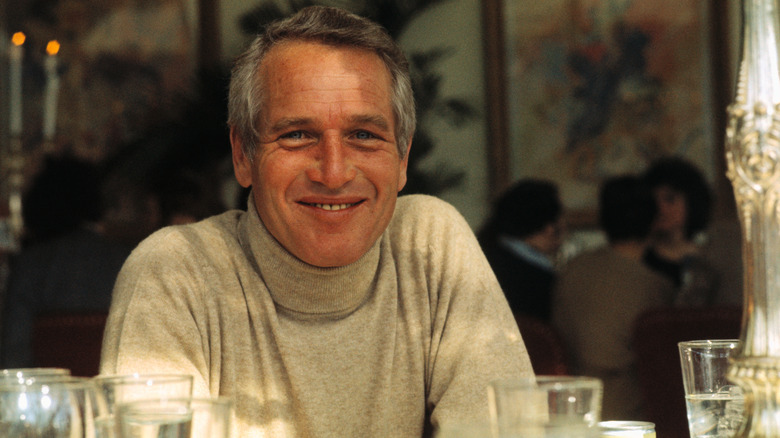 Paul Newman smiling at a restaurant table in a gray turtleneck sweater