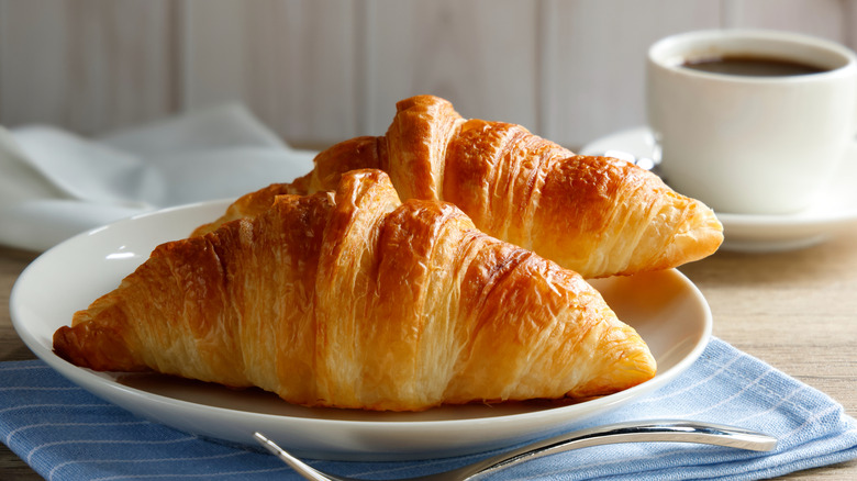 Two light and flaky croissants on a plate with a cup of coffee behind.