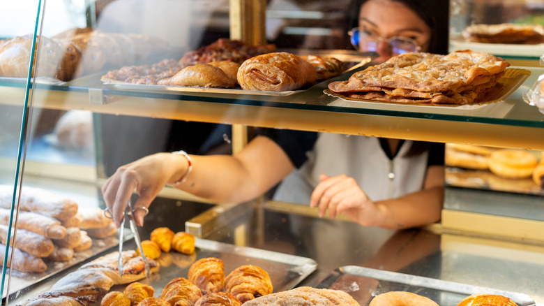An employee grabs a pastry from the display case at a bakery using tongs.