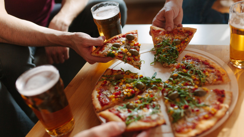 Multiple hands taking pieces of veggie pizza surrounded by full glasses of beer