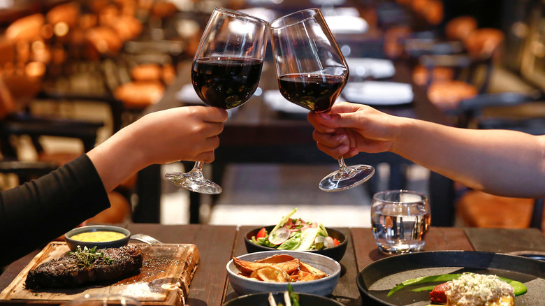 Two hands toasting with glasses of red wine and eating steak at a restaurant.