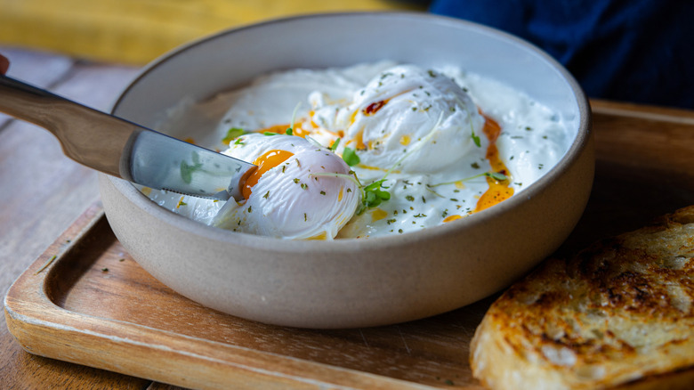 Knife cutting through one of two poached eggs in bowl on tray next to toast