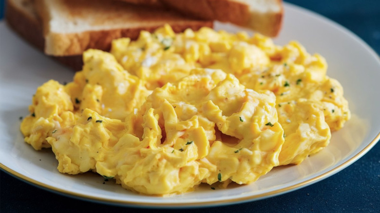 White plate with creamy scrambled eggs with toast in the background