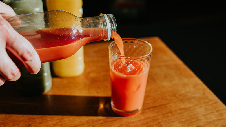 A hand pouring reddish-orange juice into a glass on a wooden table.