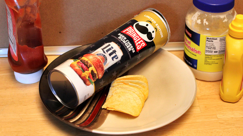 A can of the Pringles x Miller Lite beer cheese burger flavored chips on a plate surrounded by condiments that would go on a burger