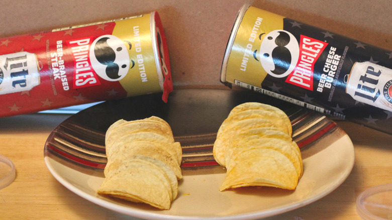 Two cans of limited edition Pringles x Miller Lite 2026 collaborative chips, beer-braised steak and beer cheese burger, beside a plate with some of the chips on it