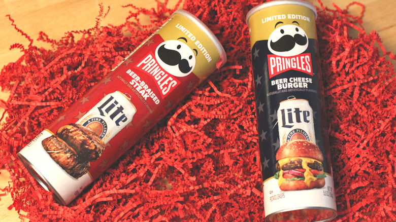 Two cans of limited edition Pringles x Miller Lite 2026 collaborative chips, beer-braised steak and beer cheese burger, on red paper
