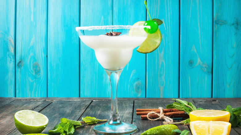 A frozen lime margarita with a green cherry and lime garnish on a wooden table with a blue background