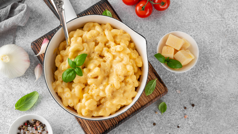 A white pot fill with Mac and cheese on a cutting board garnished with fresh basil and surrounded by other ingredients like tomatoes, pepper, and garlic.