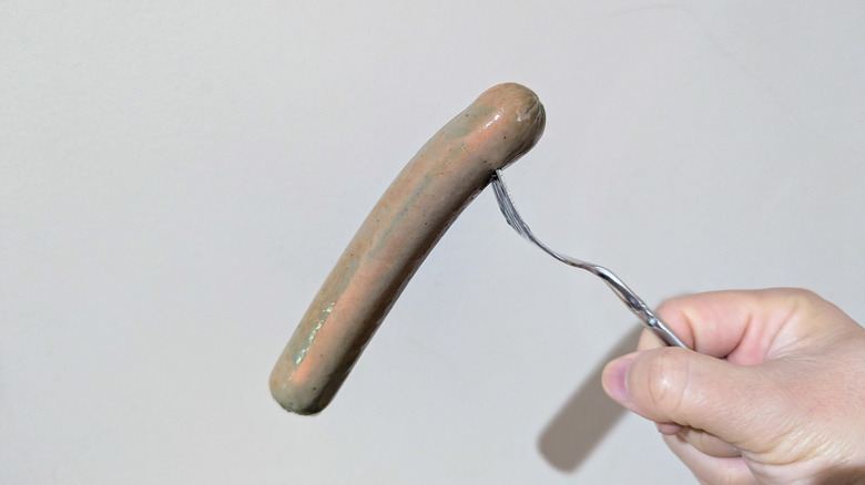 Barely blue hot dog pierced with fork held in front of wall.
